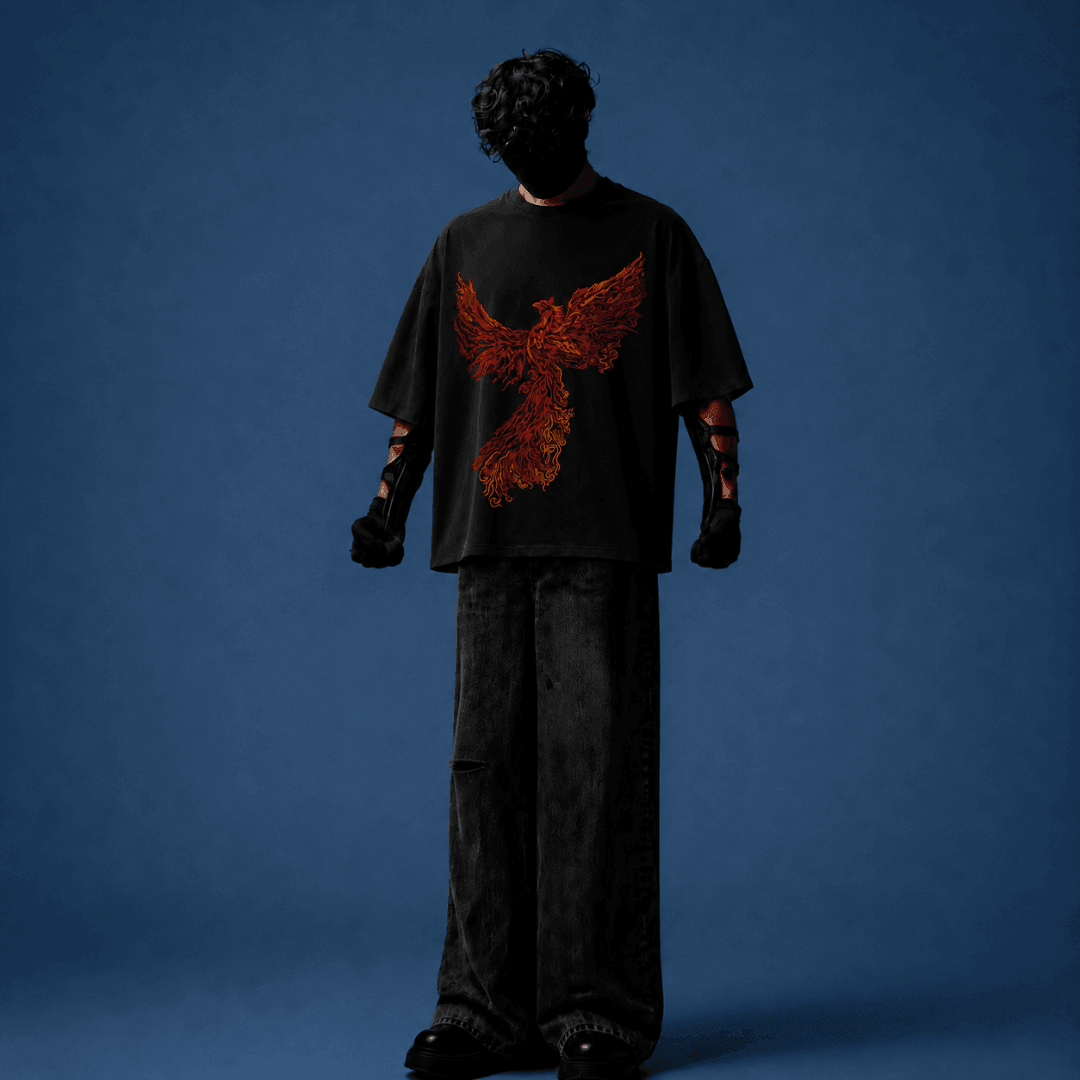 A model stands with their back to the camera wearing an oversized black t-shirt featuring a large red and orange phoenix graphic on the back, paired with dark wide-leg jeans, set against a solid blue studio background.